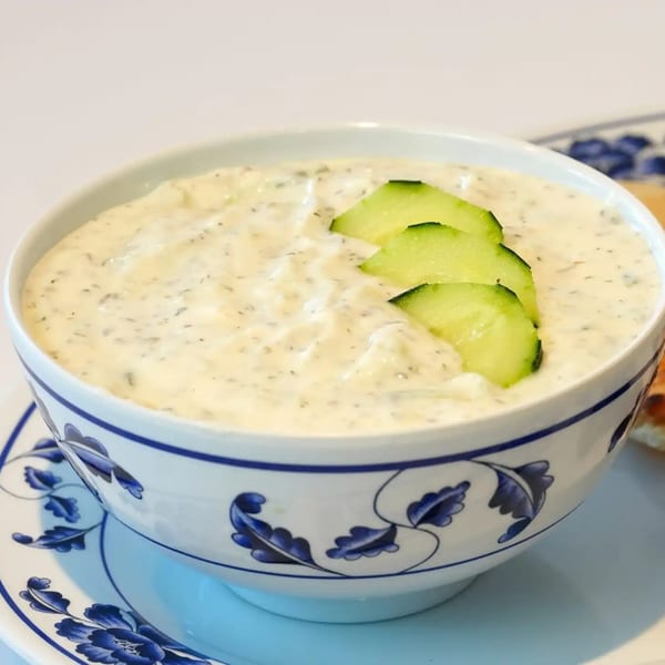 A white and blue patterned bowl filled with creamy tzatziki, garnished with three cucumber slices.