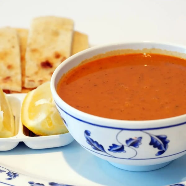A bowl of red lentil soup with lemon wedges and pita bread on a white and blue plate.